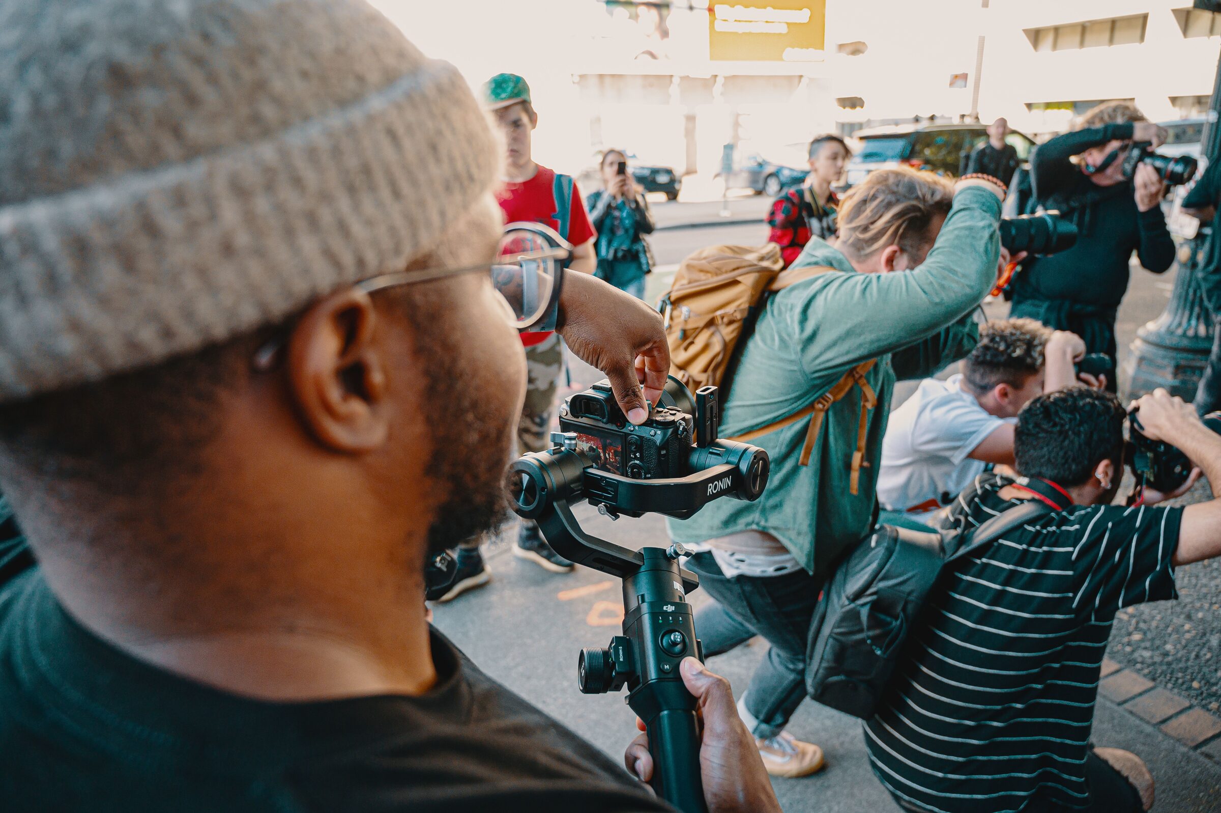 A videographer operates a gimbal-stabilized camera among a crowd of photographers at a street event.